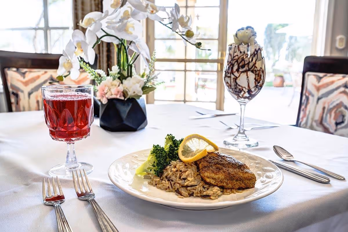A dining table set with a plate of seasoned fish, rice, and broccoli garnished with a lemon slice. There is a glass of red beverage and a dessert glass with chocolate drizzle and whipped cream. A floral centerpiece with white and pink flowers is in the background, with large windows letting in natural light.