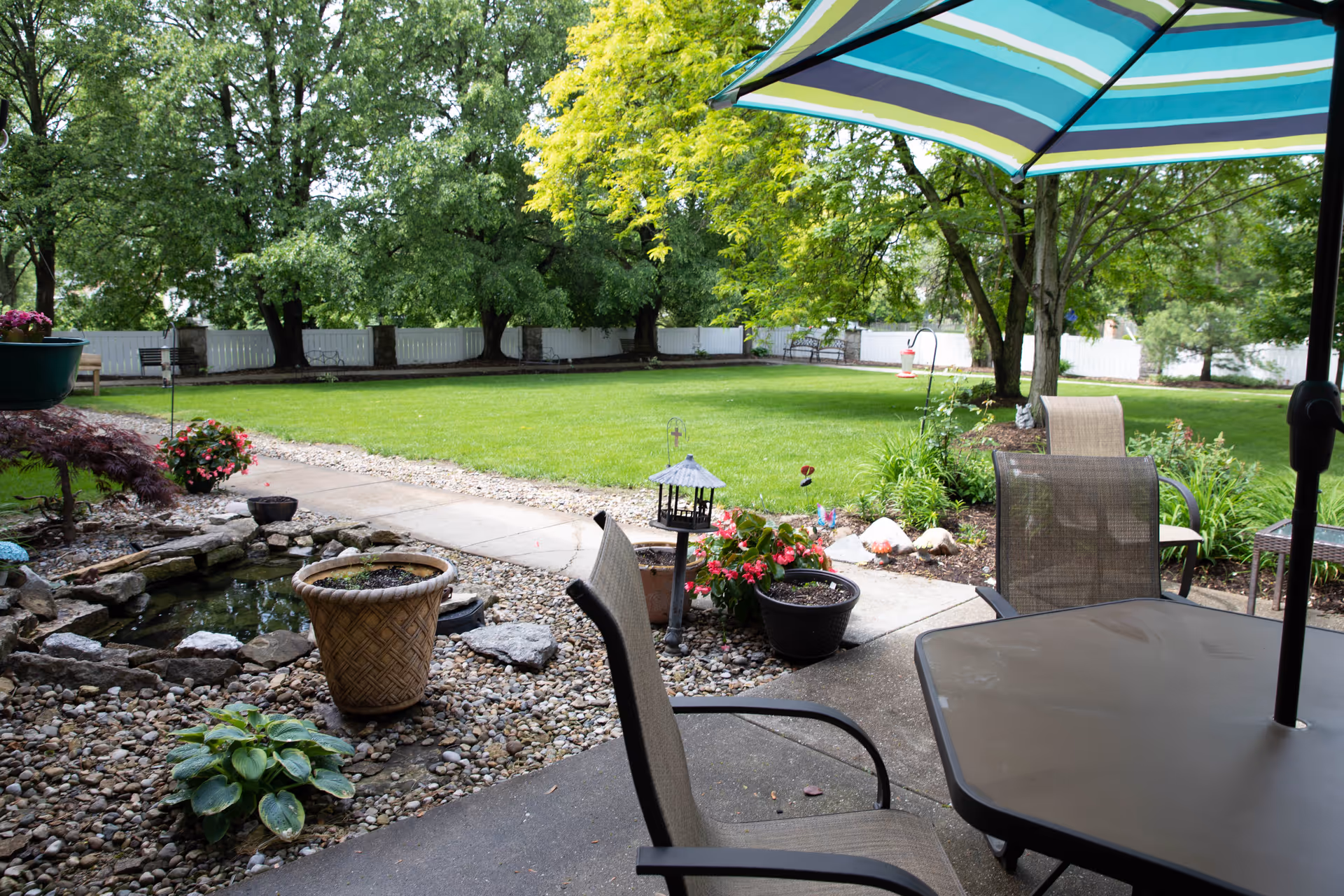 Outdoor patio area with a table and chairs under a striped umbrella, overlooking a well-maintained lawn with trees, flower pots, a small pond, and a white fence in the background.