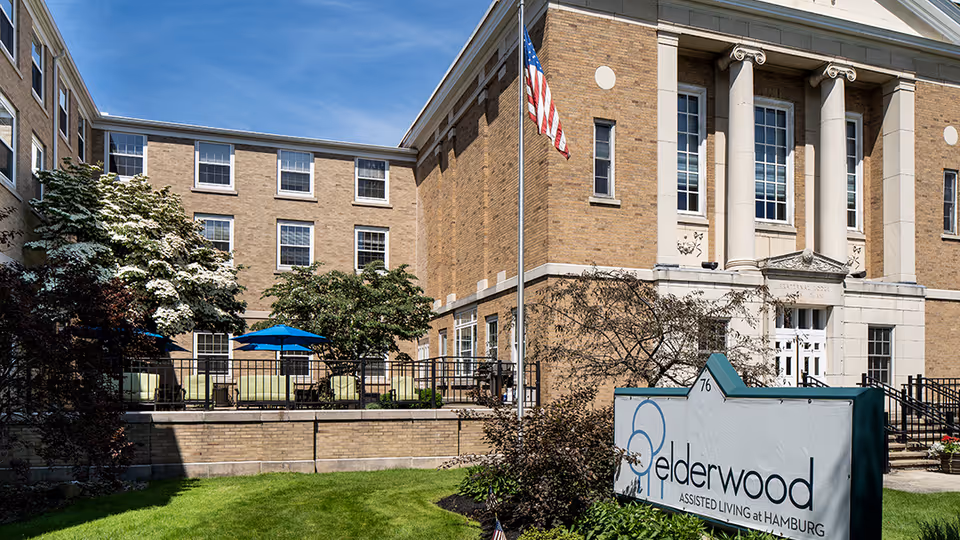Brick assisted living facility with classical columns, an outdoor patio with blue umbrellas, and a sign reading "Elderwood Assisted Living at Hamburg".