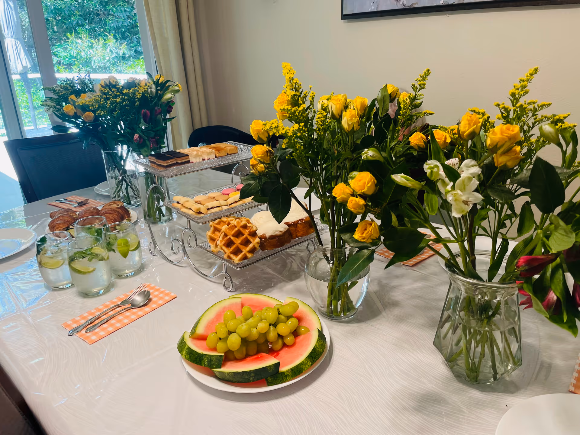 A dining table set with various refreshments including glasses of water with lime and mint, croissants, assorted pastries on a tiered stand, and a plate of watermelon slices topped with green grapes. The table is decorated with multiple vases of yellow and white flowers. The room has a large window with a view of greenery outside.