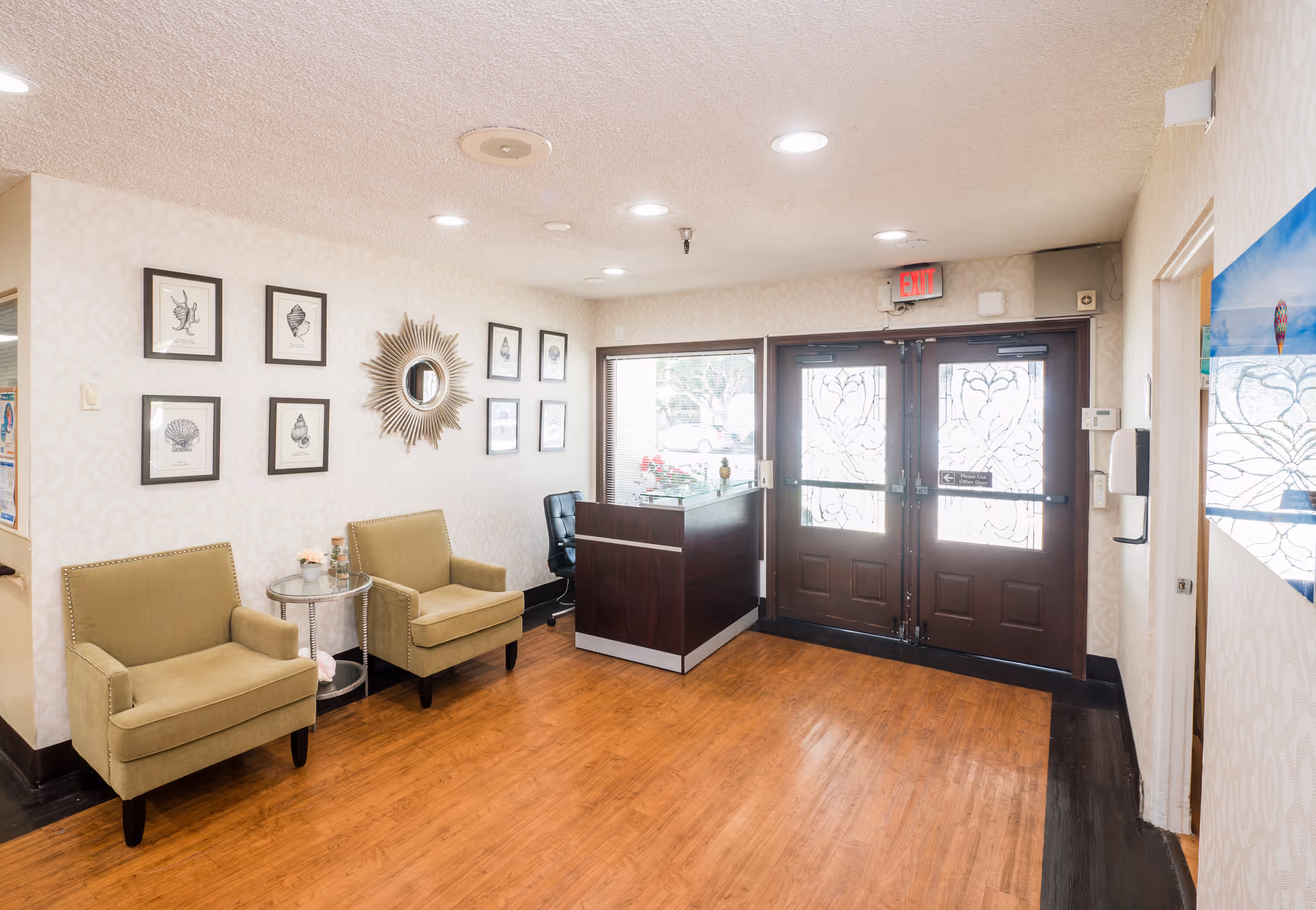 A bright and welcoming reception area with two beige armchairs and a small round glass table between them. The walls are decorated with framed shell illustrations and a sunburst mirror. There is a dark wood reception desk near double glass doors with decorative frosted patterns, and an exit sign above the doors. The floor is wooden, and the space is well-lit with ceiling lights.