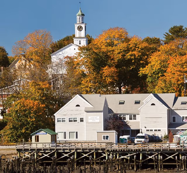 View of a waterfront building with a wooden dock in front, surrounded by trees with autumn foliage. In the background, there is a white church with a clock tower and a green spire under a clear blue sky.