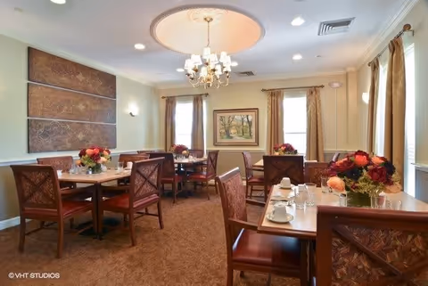 A well-lit dining room with multiple square wooden tables, each set with glasses, cups, and floral centerpieces. The room features beige walls, carpeted floor, two windows with curtains, a chandelier hanging from the ceiling, and framed artwork on the walls.