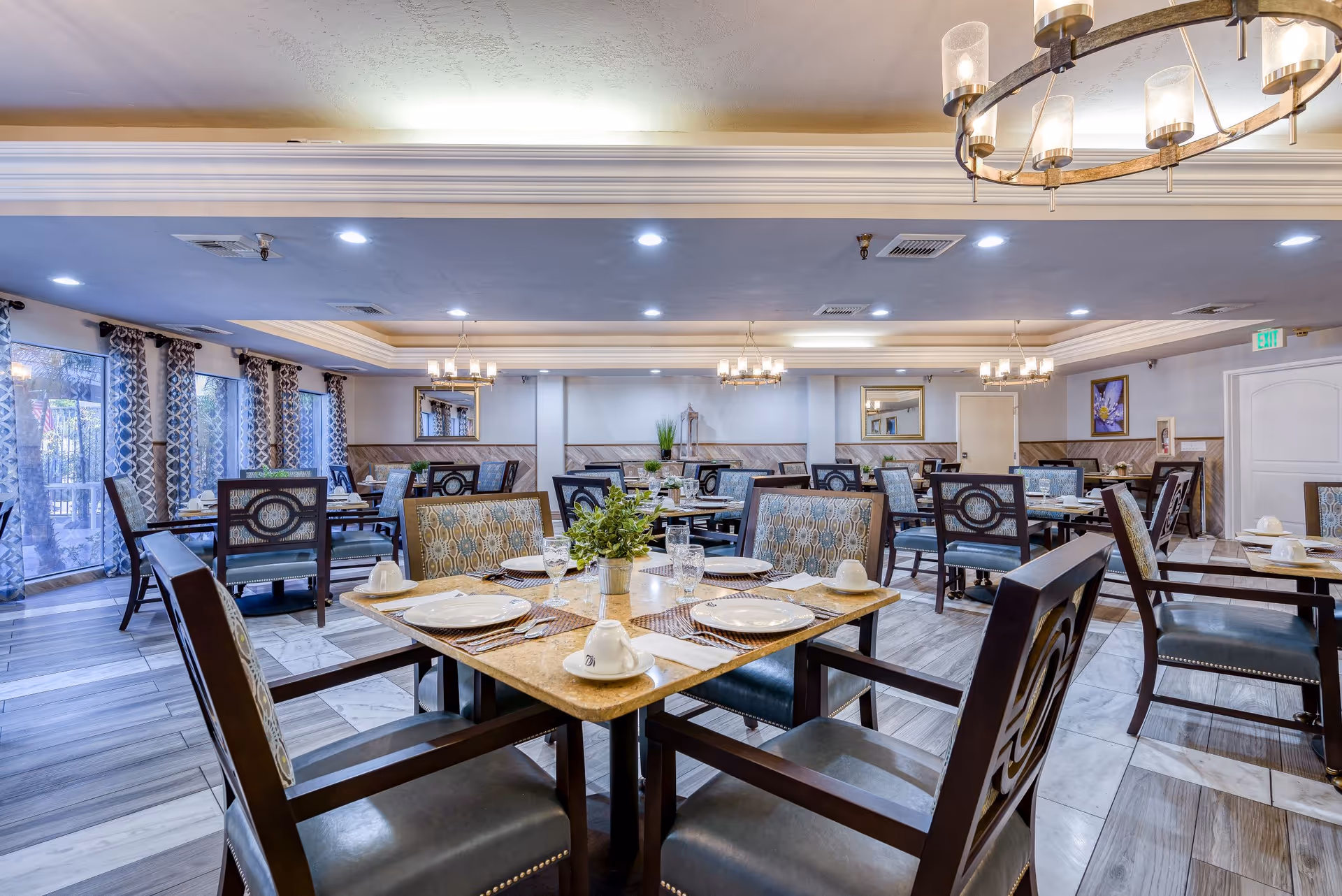 A spacious dining room in Alta Vista Senior Living with multiple tables set for meals. Each table has plates, cups, glasses, and silverware neatly arranged. The room features patterned curtains on large windows, decorative mirrors on the walls, and modern chandeliers hanging from the ceiling. The flooring is a mix of wood and tile patterns, and the overall ambiance is bright and welcoming.