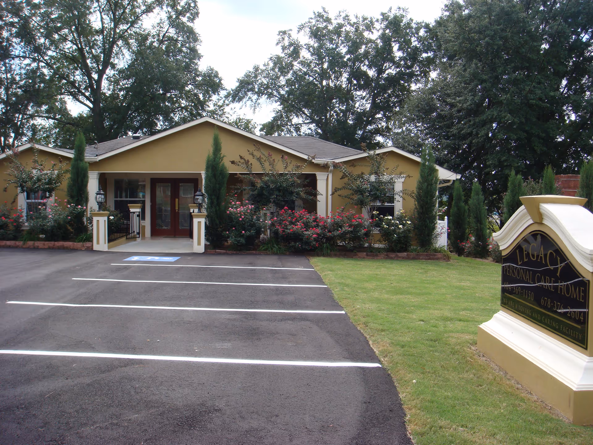 Exterior view of Legacy Personal Care Home, a single-story building with a beige facade, surrounded by trees and bushes with pink flowers. There is a paved parking lot with marked spaces and a handicap parking spot in front of the entrance. A large sign with the facility's name is visible on the right side of the image.