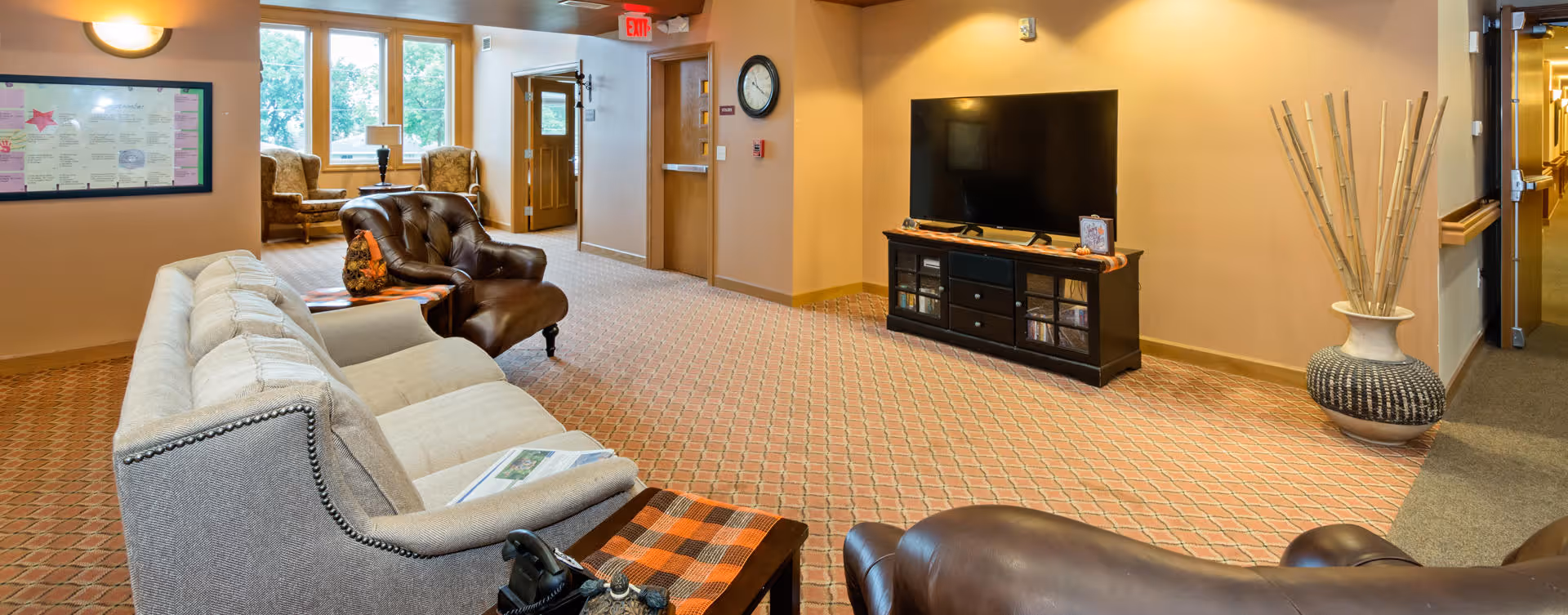 A cozy living room area in a senior living facility with a beige sofa, two brown leather armchairs, a TV on a black stand, a large vase with decorative sticks, and a window letting in natural light. The room has patterned carpet and warm beige walls.