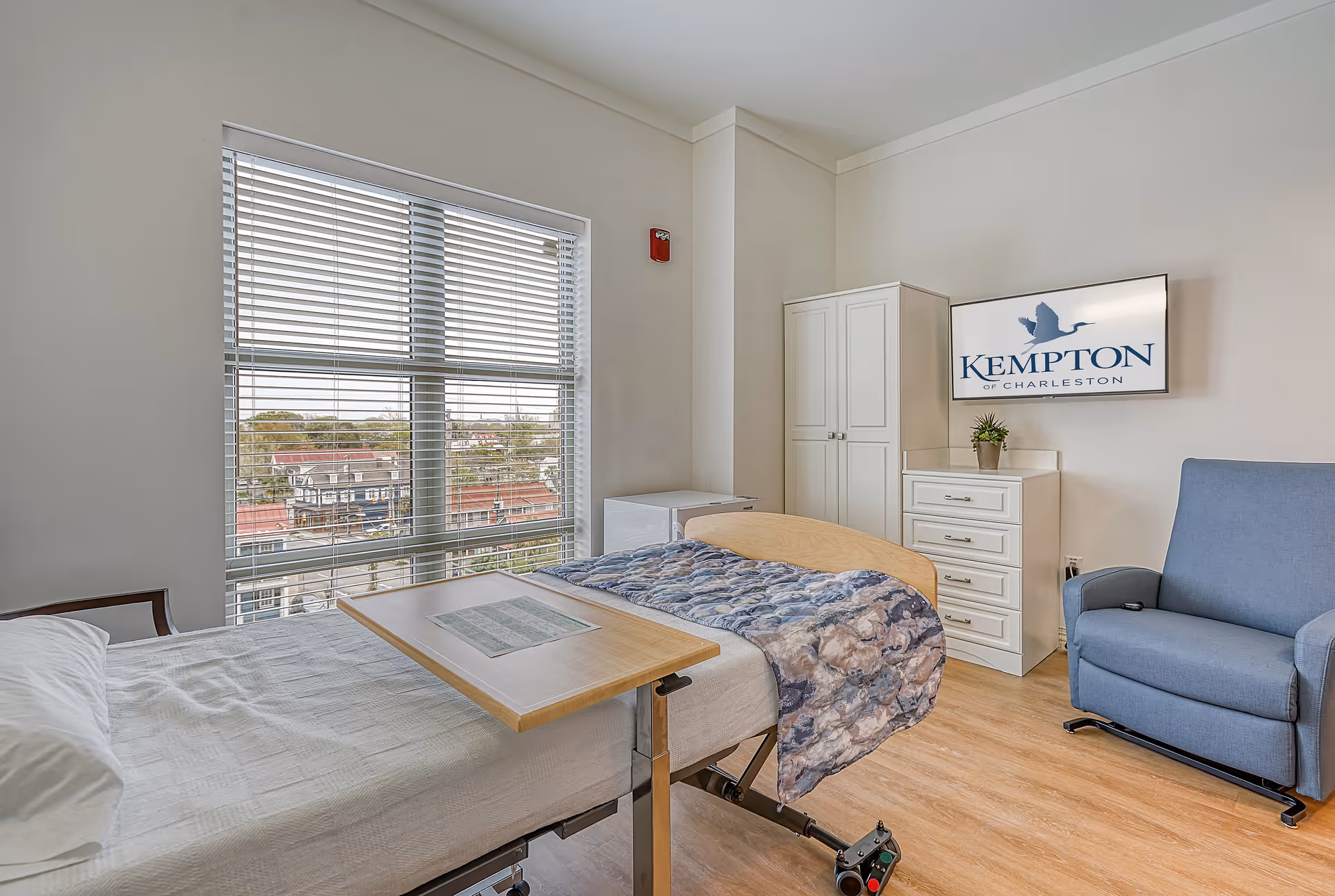Bright senior bedroom with a hospital-style bed and overbed table, armchair, dresser and wardrobe beside a large window with blinds.