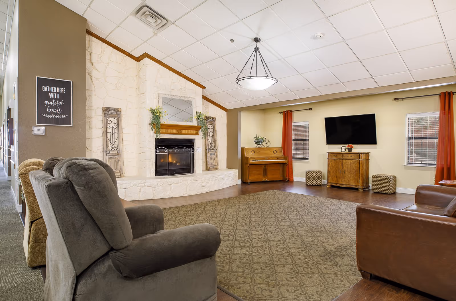 A cozy senior living facility common area with a stone fireplace, a mirror above it, and decorative plants on the mantel. There are several comfortable chairs and a leather sofa arranged around a large patterned rug. A wooden piano is placed against the far wall between two windows with red curtains. A flat-screen TV is mounted on the wall above a wooden cabinet, flanked by two small patterned ottomans. The ceiling has white tiles with a hanging light fixture.