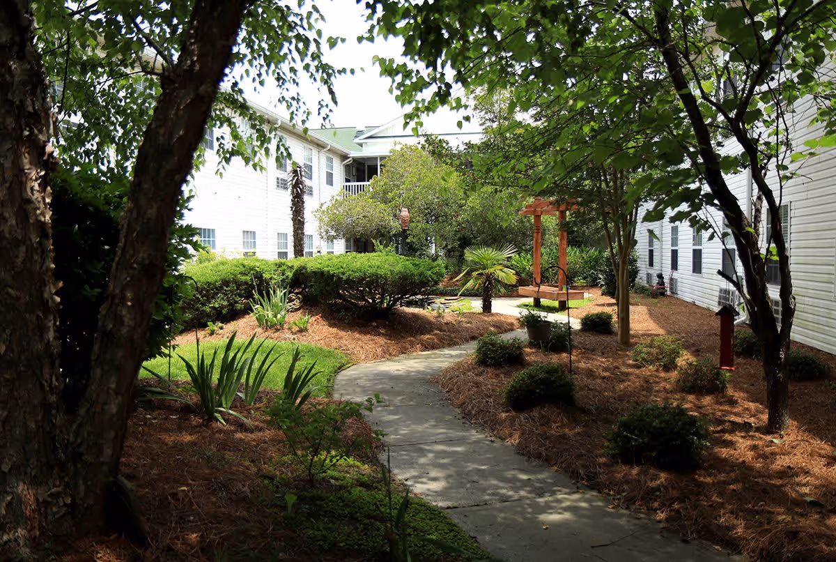 A landscaped outdoor garden area with a winding concrete pathway surrounded by green bushes, trees, and plants. The garden is situated between white buildings with multiple windows, and there is a wooden swing set visible in the background.