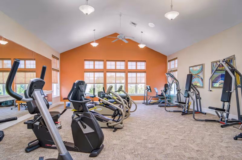 A spacious fitness room with various exercise equipment including stationary bikes, elliptical machines, and weight machines. The room has large windows with natural light, an orange accent wall, beige walls, carpeted floor, ceiling fans, and framed artwork on the walls.