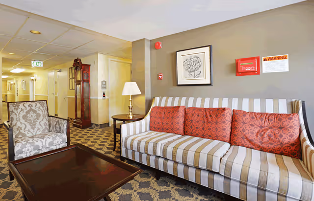 A senior living lounge area with a striped sofa with red cushions, a patterned armchair, coffee table, lamp, and a hallway in the background.