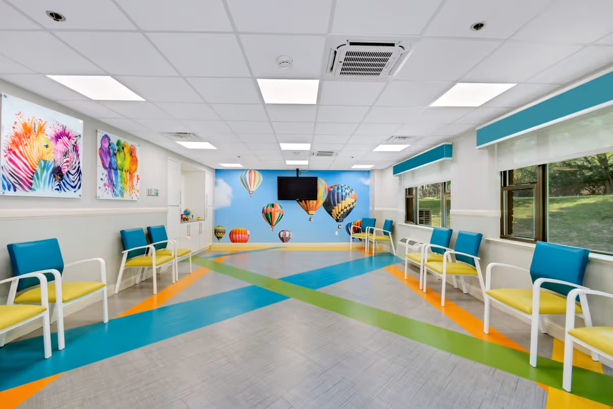 A brightly lit room with colorful chairs arranged along the walls. The floor has vibrant blue, green, and orange stripes. The far wall features a mural of hot air balloons floating in a blue sky with clouds. There are large windows on one side letting in natural light, and two colorful animal paintings on the opposite wall.
