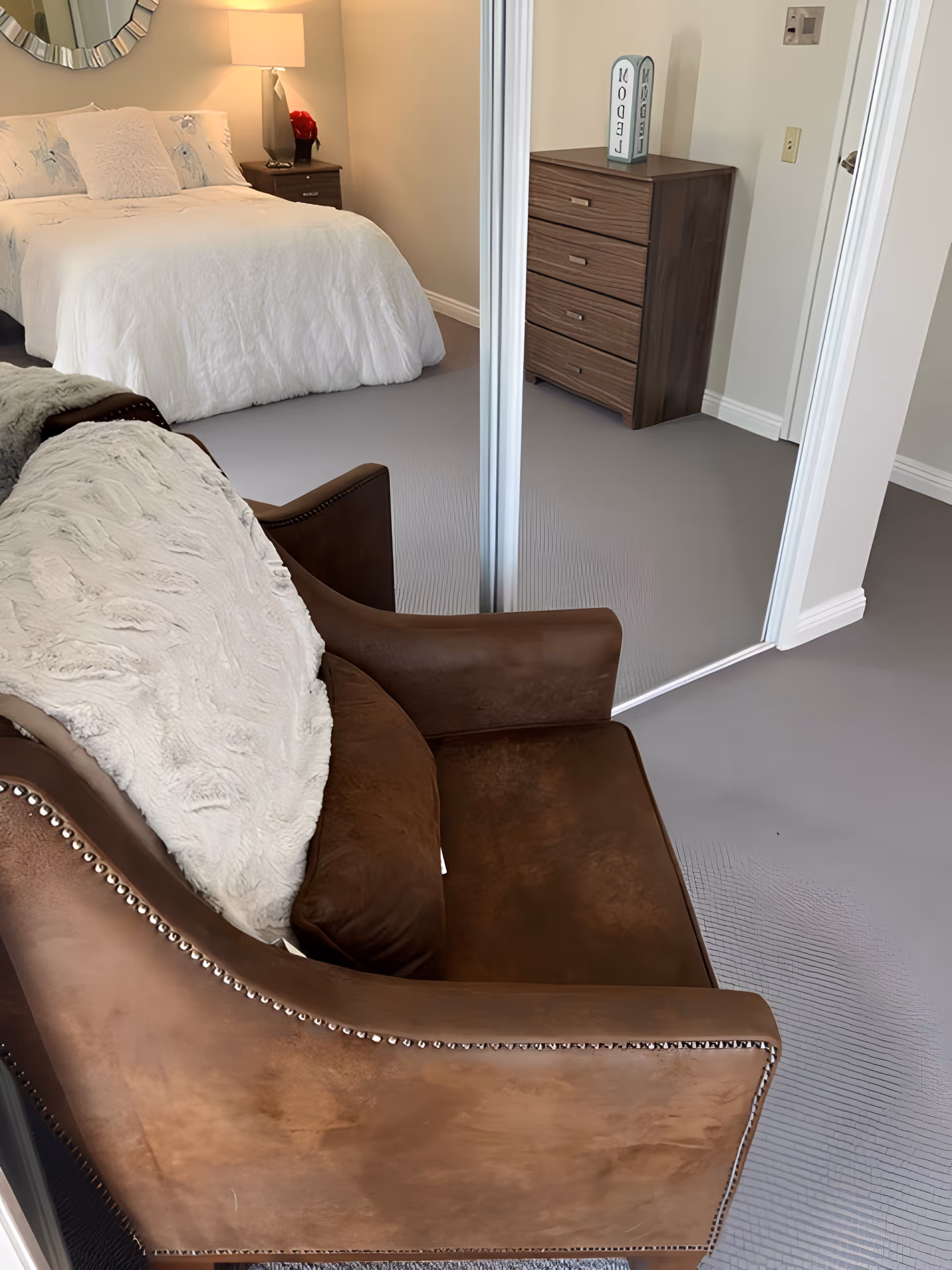 Cozy bedroom with a brown leather armchair in the foreground, a white bed and lamp in the background, and mirrored closet doors reflecting a dresser.