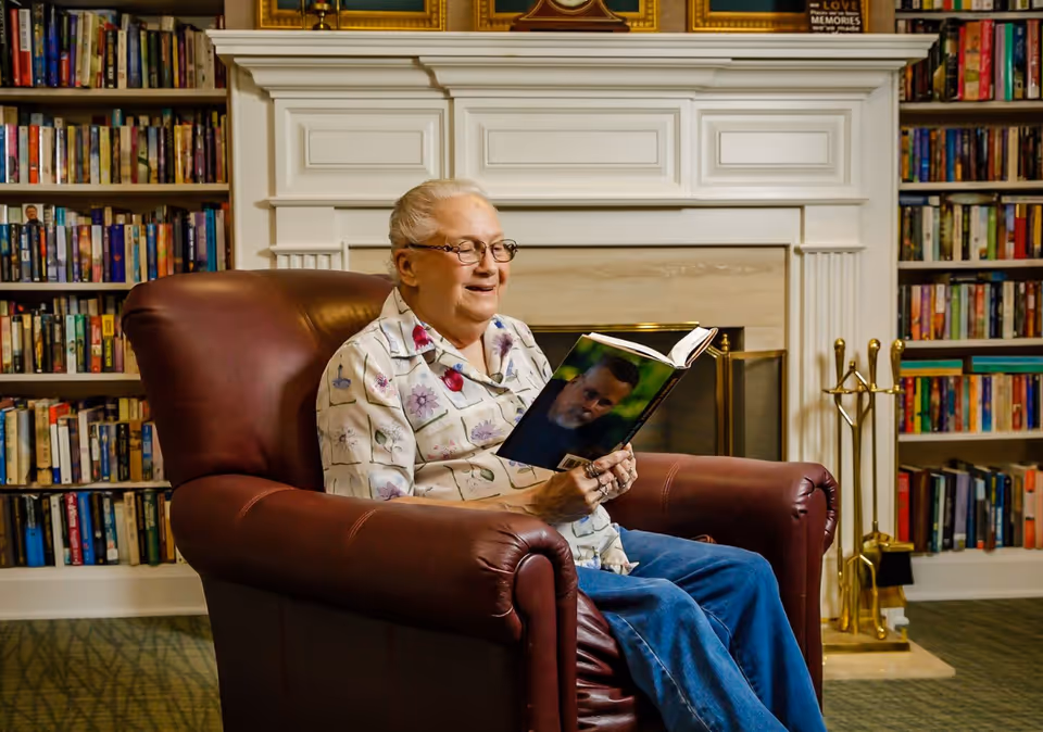 An elderly woman sits in a leather armchair reading a book in a room with bookshelves and a fireplace.