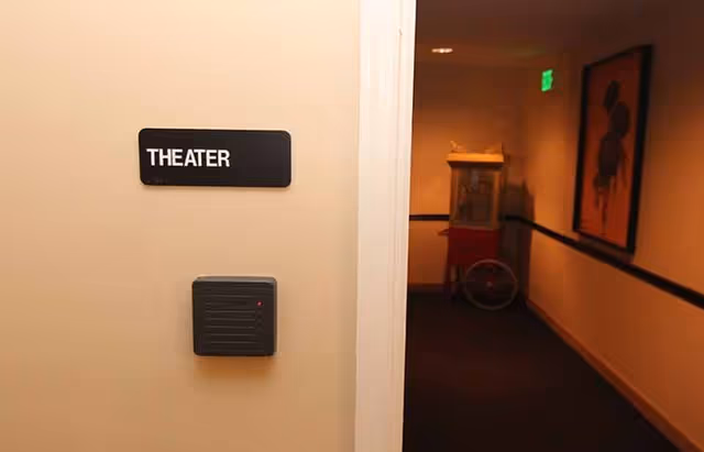 A hallway with a sign on the wall labeled 'THEATER' next to a card reader device. In the background, there is a popcorn machine and a framed picture on the wall under warm lighting.