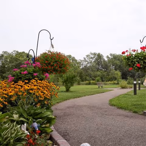 A winding paved garden path through a grassy park with hanging flower baskets and a bench in the distance.