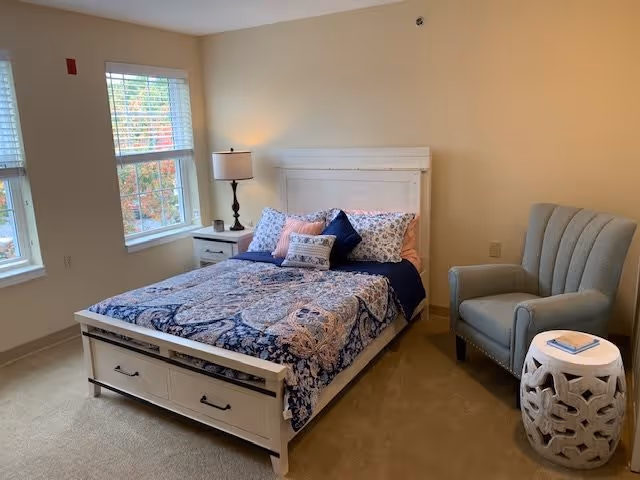 A cozy bedroom with a white wooden bed frame featuring drawers underneath, a bed made with patterned blue and pink bedding and multiple pillows. There is a white nightstand with a lamp next to the bed, two windows with blinds letting in natural light, a gray upholstered armchair, and a white decorative side table with a book on it. The walls are painted a light beige color.