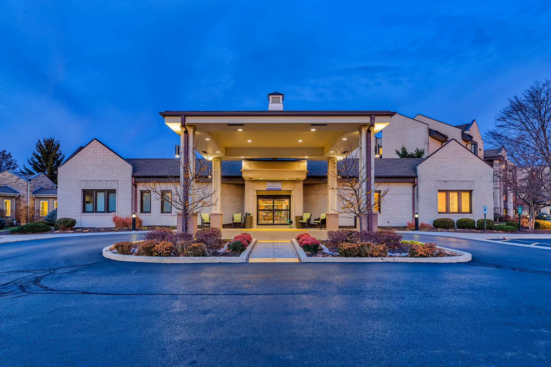 Exterior front view of Harbour House facility at dusk with a covered entrance, landscaped flower beds, and illuminated windows.