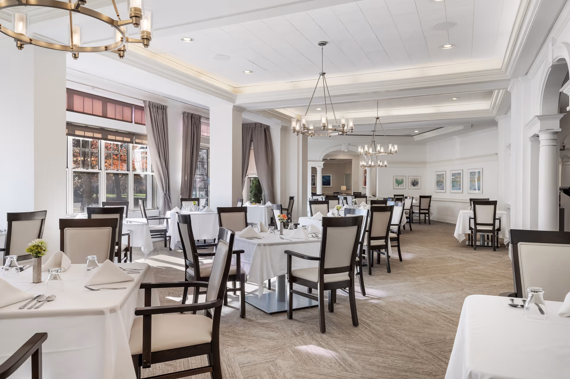 Spacious bright dining room with white-clothed tables and dark wooden chairs, chandeliers, and large windows.
