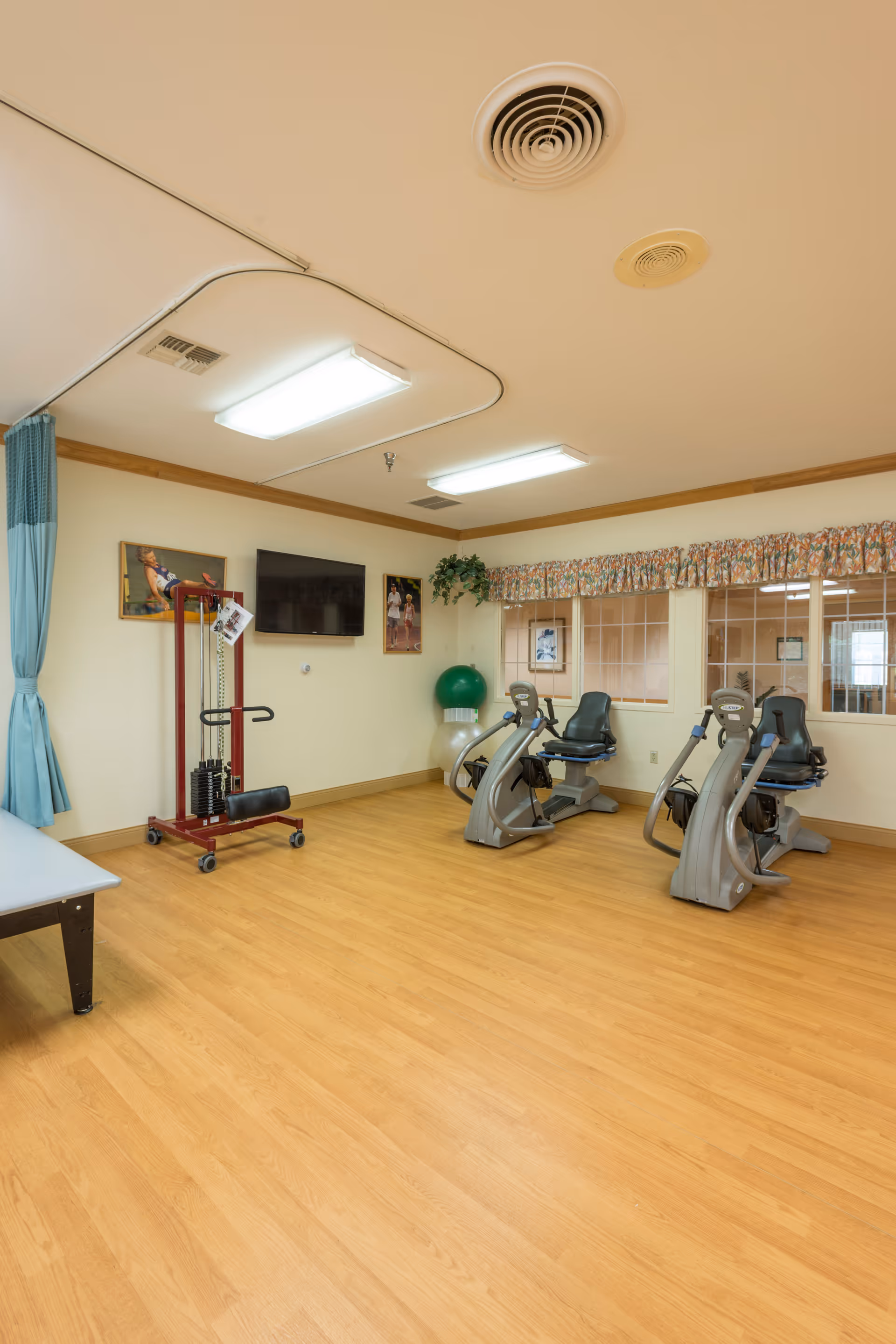 A small exercise room with wooden flooring, two recumbent exercise bikes, a red weight machine, a green exercise ball, and a flat-screen TV mounted on the wall. The room has cream-colored walls, a window with floral valance curtains, and framed pictures on the walls.