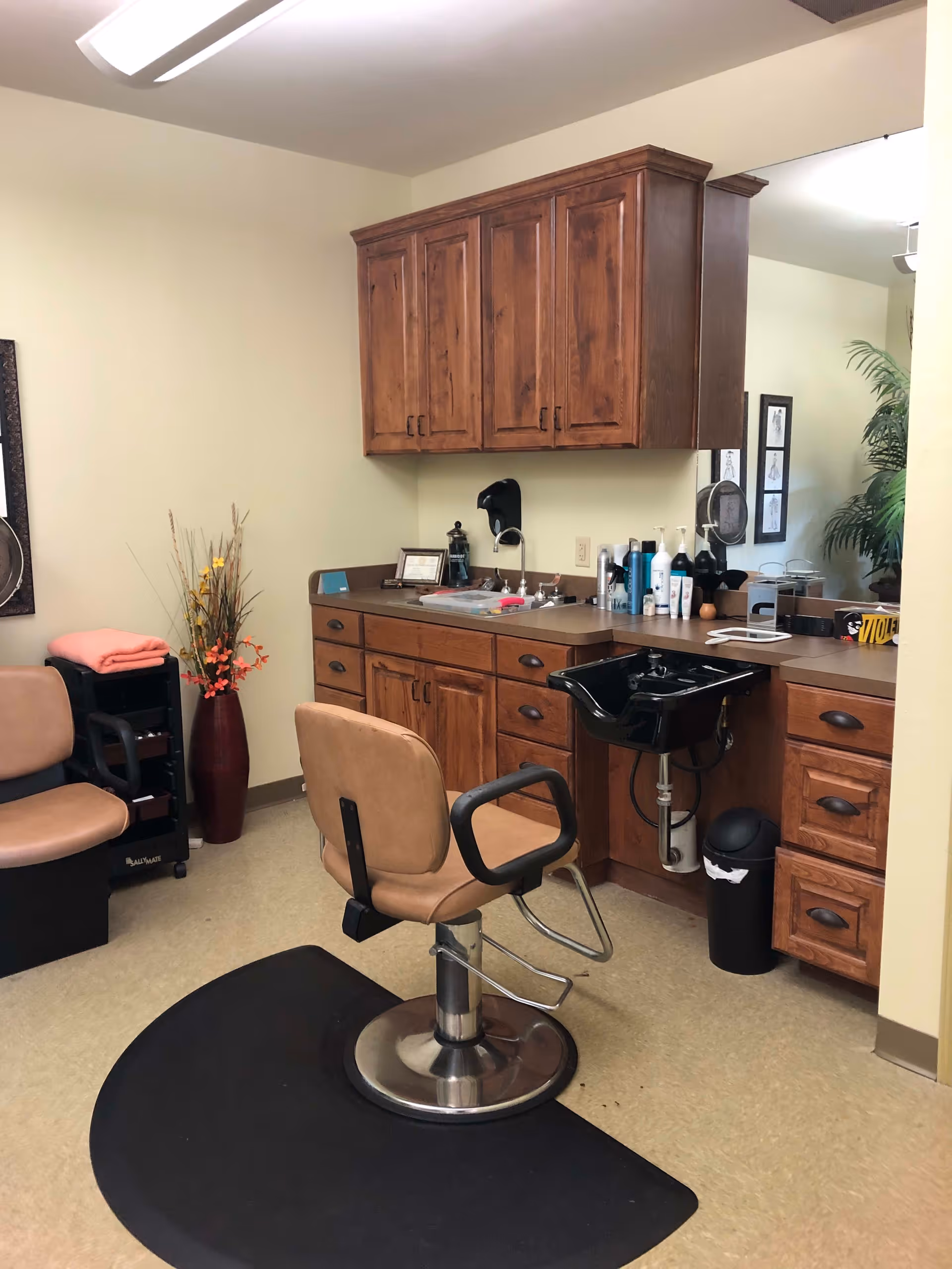 Interior of a hair salon area in a senior living facility with a salon chair on a black mat, wooden cabinets, a black wash basin, various hair care products on the counter, a large mirror, a small trash can, a vase with decorative flowers, and another chair in the corner.