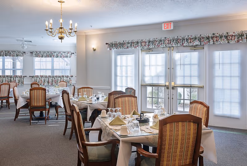 A bright dining room with several tables set for a meal, featuring wooden chairs with patterned upholstery, white tablecloths, folded napkins, glassware, and cups. The room has large windows with floral valances and double glass doors letting in natural light. A chandelier hangs from the ceiling.
