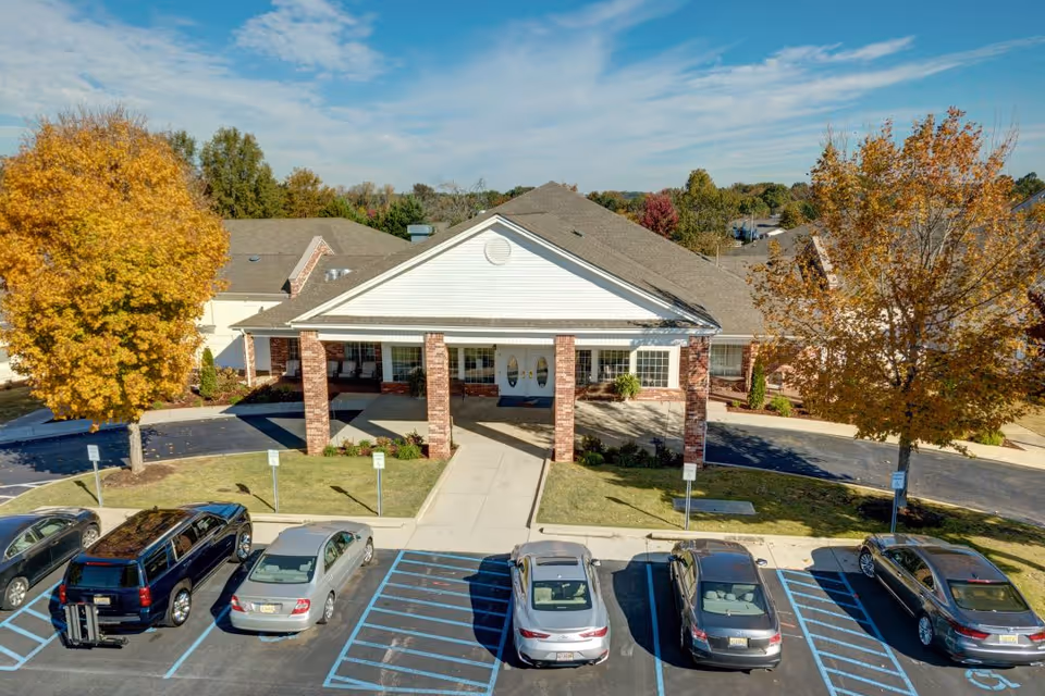 Front entrance of a single-story senior living facility with a covered portico supported by brick columns, several parked cars, and autumn trees.