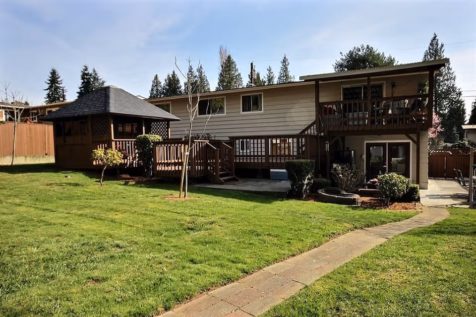Backyard of a two-story house with a wooden deck, gazebo, lawn and paved walkway.