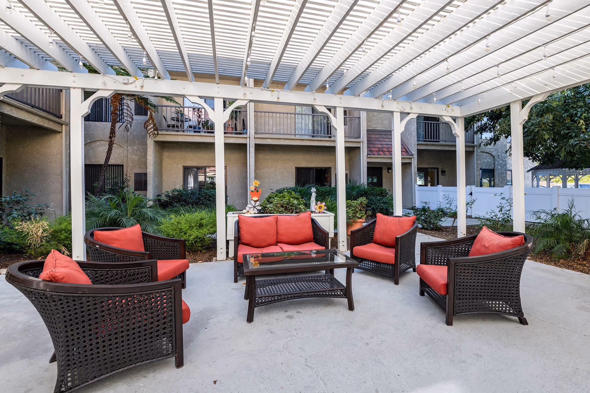 Outdoor patio area with a white pergola overhead, featuring dark wicker furniture with red cushions arranged around a glass-top coffee table. The patio is surrounded by greenery and the exterior walls of a building with balconies and windows.