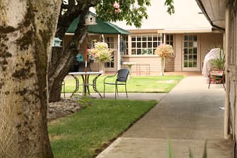 Outdoor courtyard area with a tree in the foreground, a small round table with two chairs on a grassy patch, and a building with large windows and doors in the background.