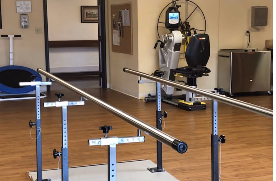 Indoor rehabilitation room with parallel walking bars and a seated exercise machine on wood floors.