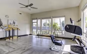 Bright interior fitness room with large windows, a ceiling fan, and exercise equipment including a stationary bike.