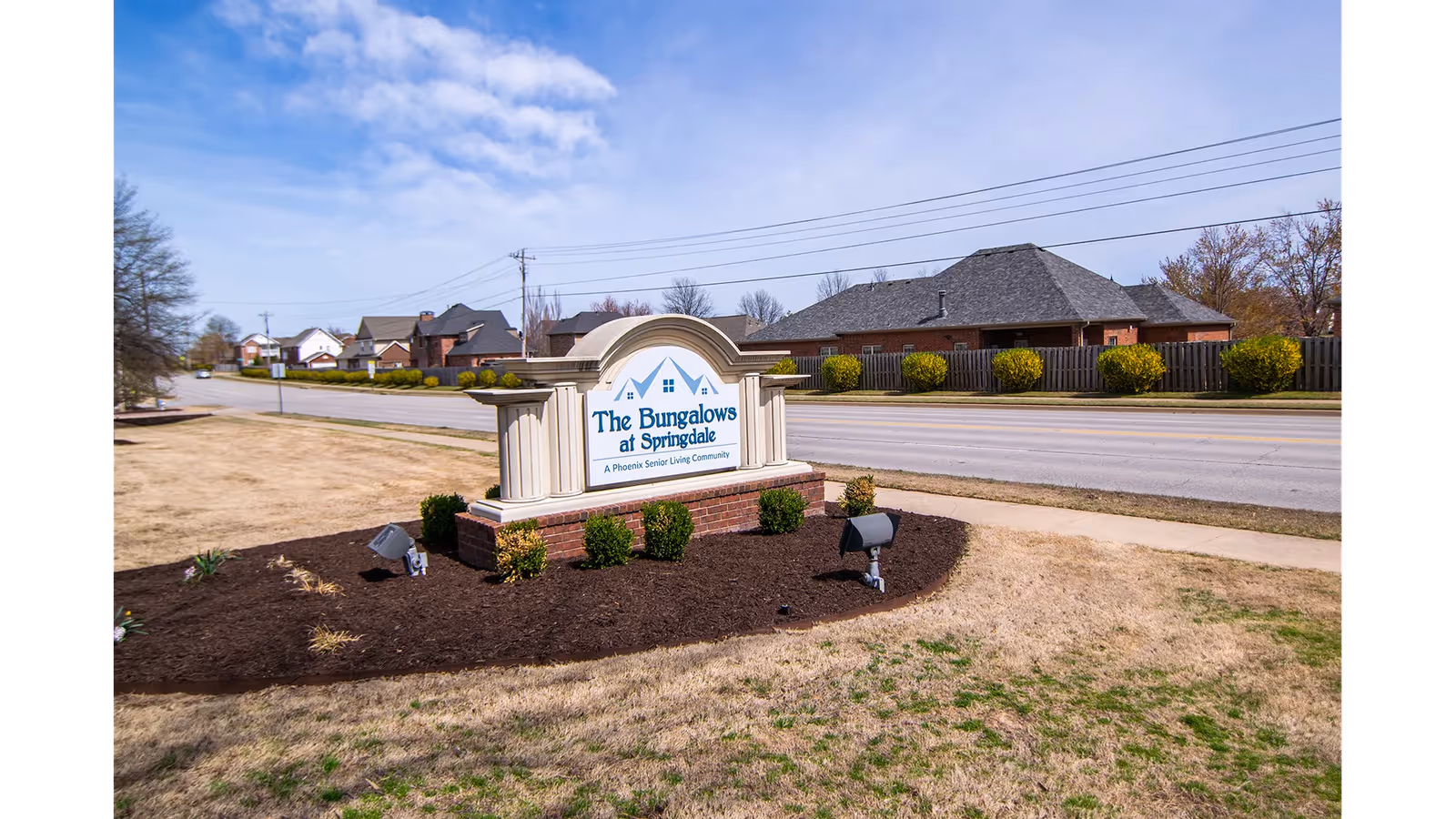 Outdoor view of a street with a landscaped area featuring a sign that reads 'The Bungalows at Springdale, A Phoenix Senior Living Community'. The sign is surrounded by mulch and small bushes, with houses and a wooden fence visible across the street under a partly cloudy sky.