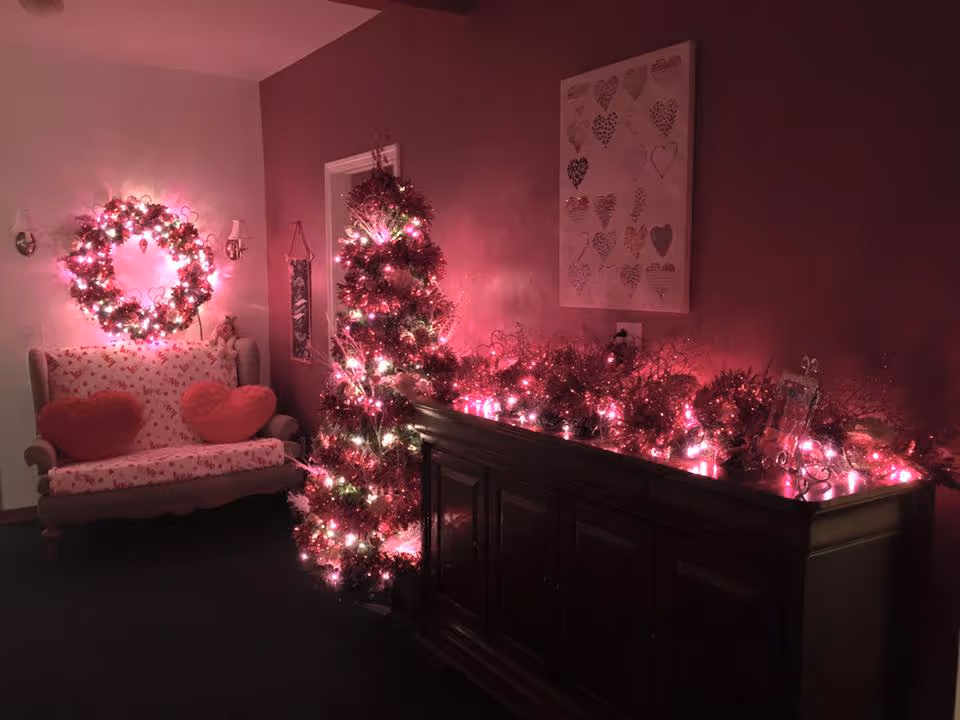 A cozy room decorated with pink and red lights for Valentine's Day, featuring a small Christmas tree with pink lights, a wreath with pink lights on the wall, a loveseat with heart-shaped pillows, and a wooden cabinet adorned with pink light decorations and heart-themed artwork on the wall.