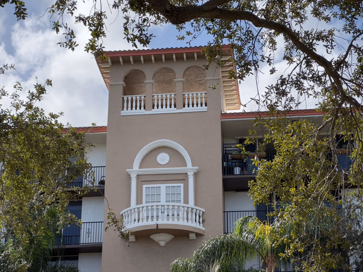 Front facade of a multi-story beige stucco building with a decorative arched balcony tower and surrounding trees.