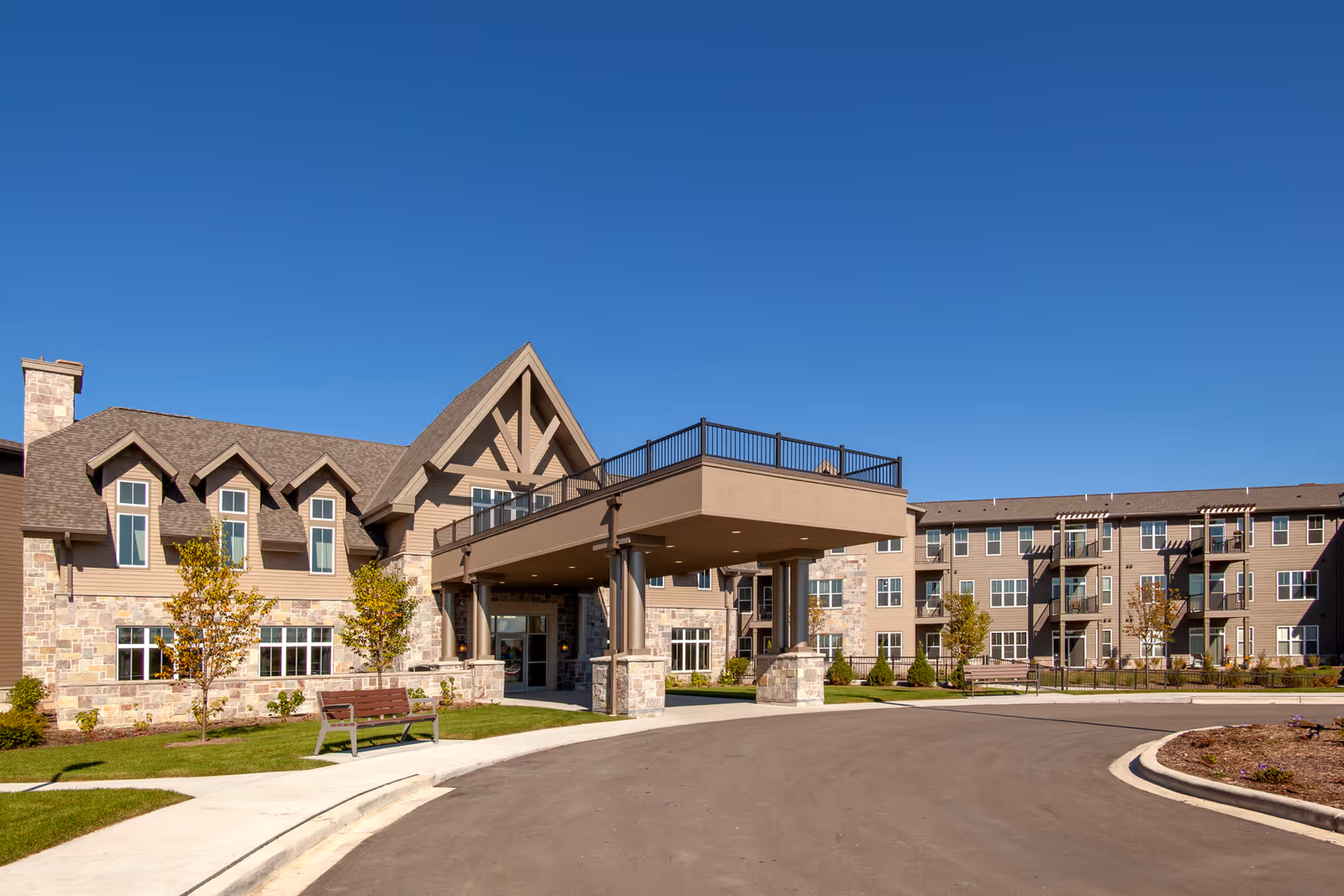 Exterior view of a senior living facility named Fairway Knoll, featuring a large building with stone and beige siding, multiple windows, a covered entrance supported by columns, landscaped greenery, benches, and a clear blue sky.