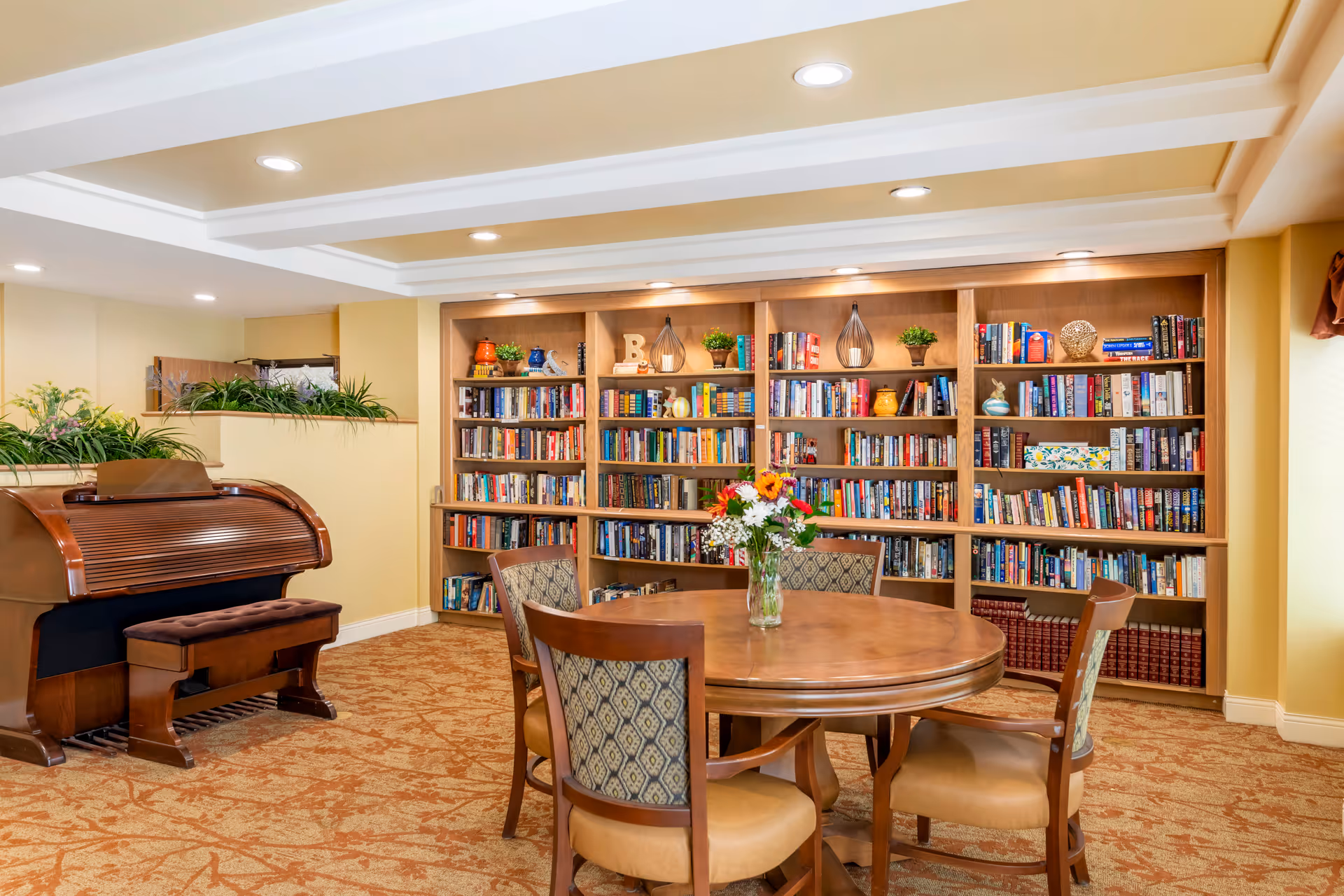 A cozy interior room featuring a round wooden table with four cushioned chairs around it. On the table is a glass vase with a colorful bouquet of flowers. Behind the table is a large wooden bookshelf filled with books and decorative items. To the left, there is a wooden roll-top piano with a matching bench. The room has warm yellow walls, recessed lighting, and a patterned carpet.