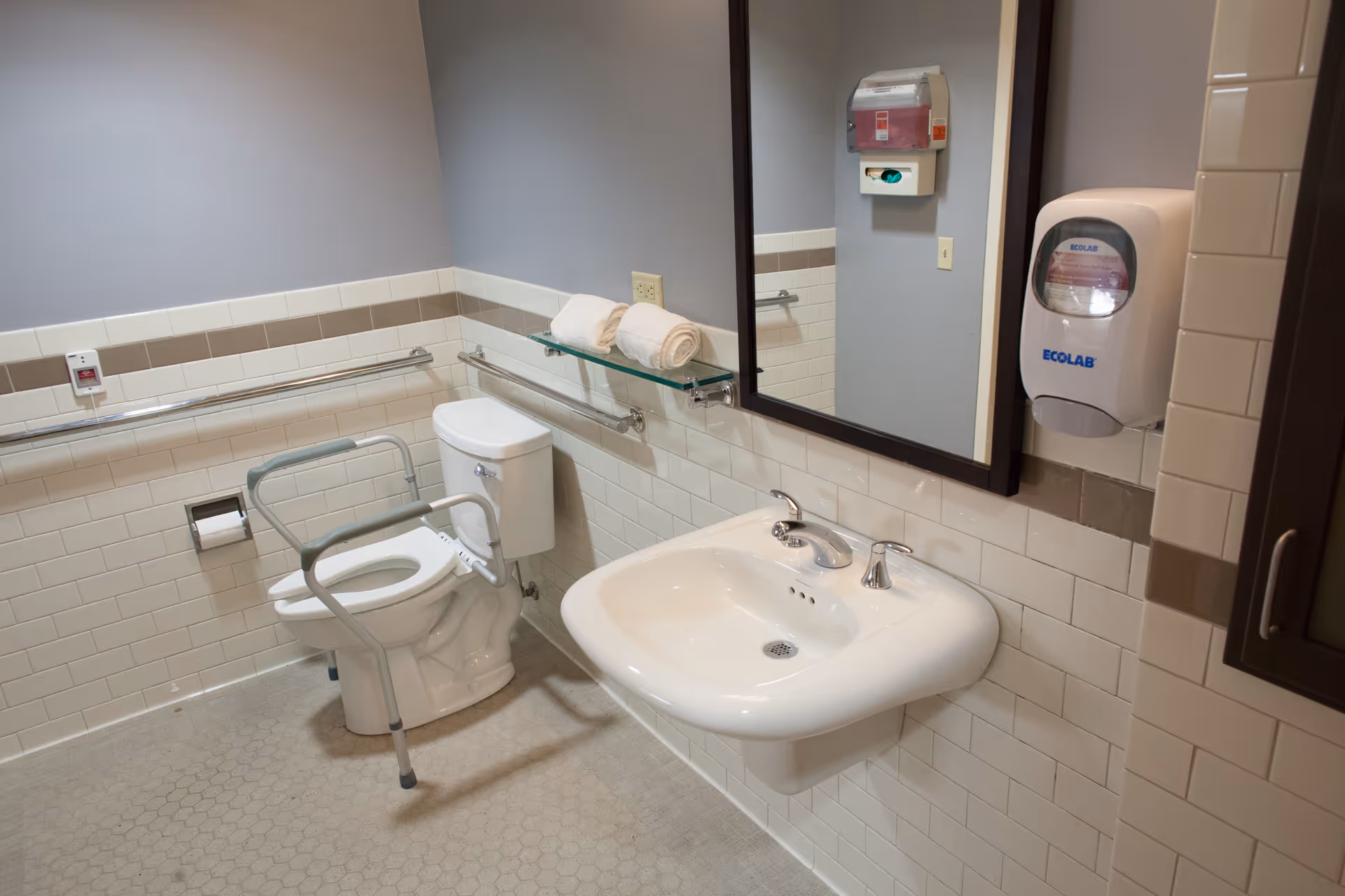 A clean, accessible bathroom with a white toilet equipped with safety rails, a white sink with two faucets, a large mirror above the sink, a glass shelf holding two rolled white towels, and a wall-mounted soap dispenser labeled ECOLAB.