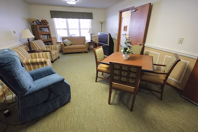 A cozy living room area with a blue recliner, two striped armchairs, a beige loveseat, a wooden table with four chairs, a TV on a stand, a bookshelf, and a window with a green valance letting in natural light.