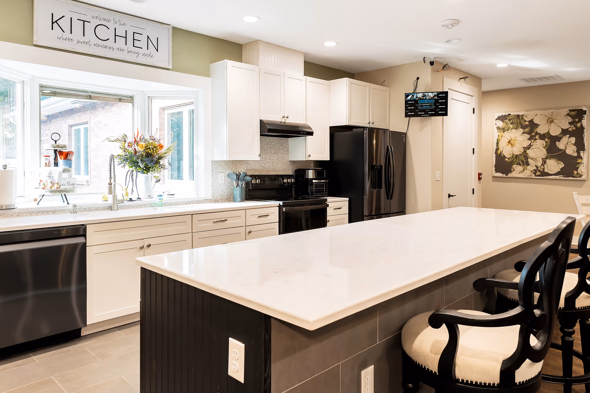 Bright and modern kitchen with white cabinets, stainless steel appliances including a dishwasher, stove, and refrigerator. A large white marble island with black cushioned chairs is in the foreground. A window above the sink lets in natural light, and a sign above the window reads 'welcome to the KITCHEN where sweet memories are being made.' There is a floral painting on the wall and a small mounted TV screen near the ceiling.