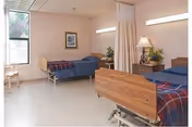 A clean and well-lit room with two hospital-style beds, each with a wooden headboard and footboard, covered with blue bedding and red plaid blankets. A small wooden nightstand with a lamp and a plant is positioned between the beds. A window with a view of greenery is on the left wall, and a framed picture hangs above one of the beds. A privacy curtain is partially drawn between the beds.