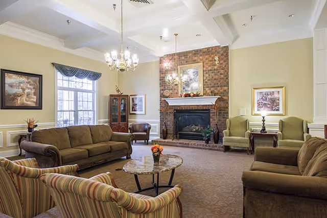 A cozy living room with a brick fireplace as the focal point, surrounded by various seating options including a brown sofa, two striped armchairs, two green armchairs, and a brown armchair. The room features a chandelier, framed artwork on the walls, a window with a valance, and a small table with a flower arrangement in the center.