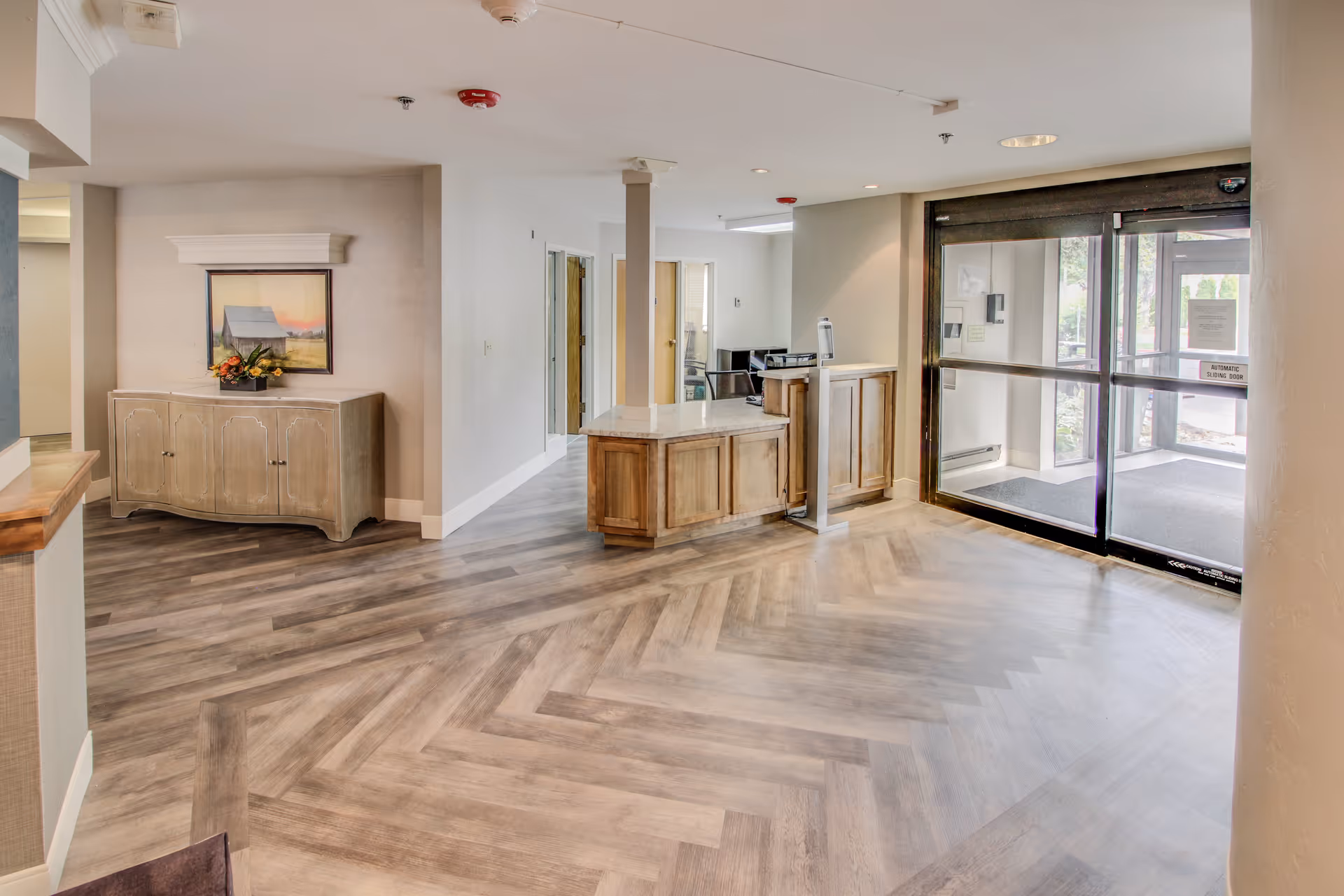 Interior view of a senior living facility lobby area with a wooden reception desk, a cabinet with a framed picture and flower arrangement on top, and glass automatic sliding doors leading outside.