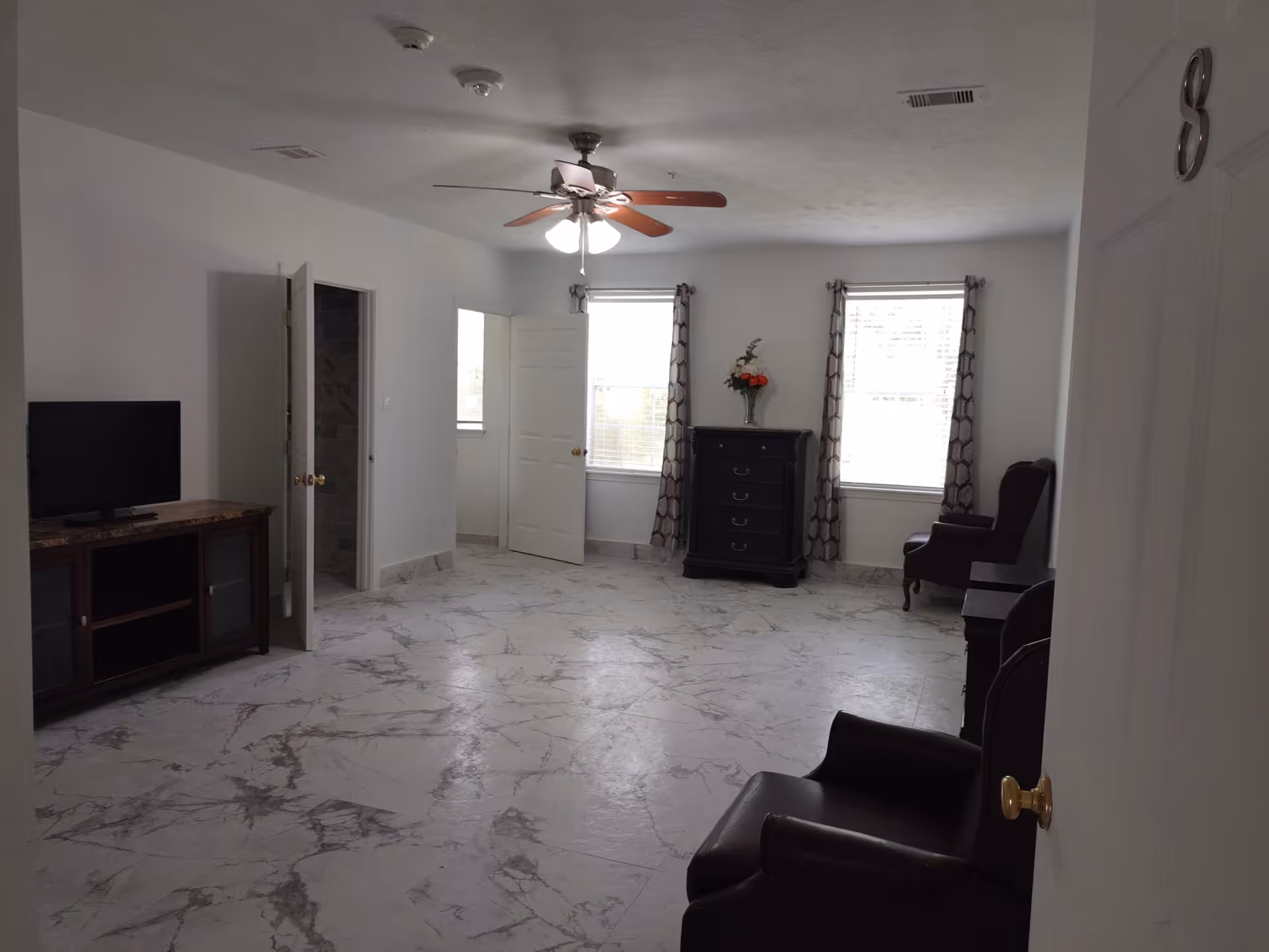 Spacious living room with marble-patterned floor, ceiling fan, TV cabinet, armchairs, and a dresser between two windows.