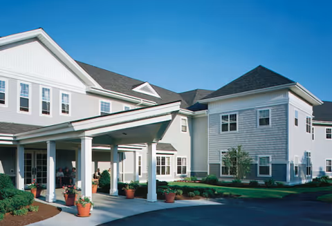 Exterior view of a senior living facility building with white siding and multiple windows under a clear blue sky. The entrance has a covered driveway supported by white columns, with potted plants placed around the entrance area.