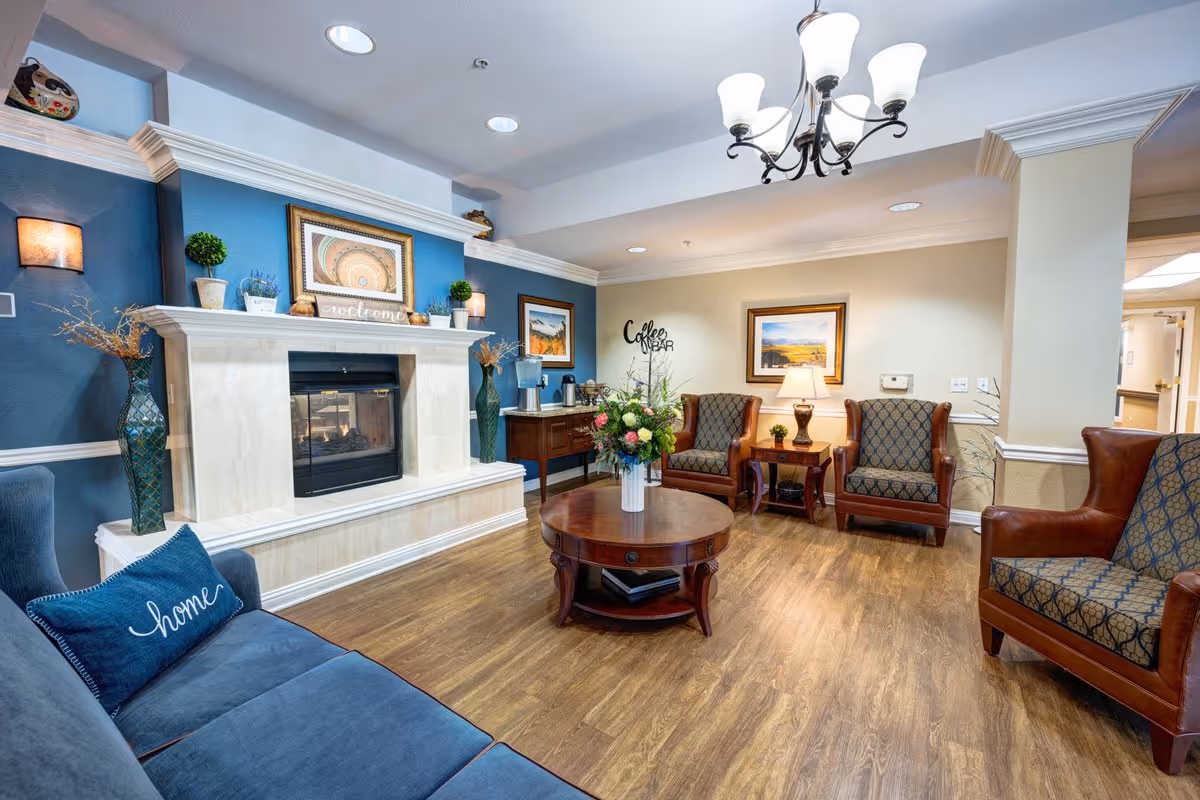 Cozy senior living common room featuring a fireplace, armchairs and sofa arranged around a central coffee table with a floral arrangement.