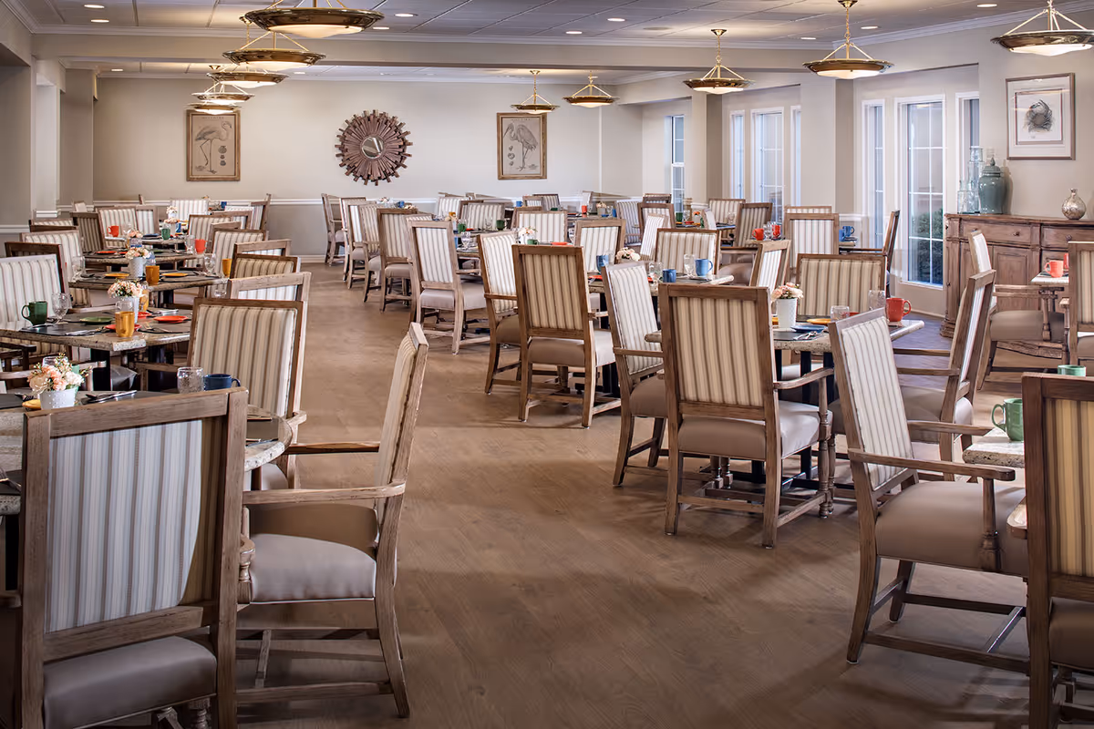 A spacious dining room in a senior living facility with multiple wooden tables and striped cushioned chairs arranged neatly. Each table is set with colorful cups, plates, and small flower arrangements. The room has large windows and glass doors letting in natural light, with framed artwork and decorative mirrors on the walls. Ceiling lights hang evenly spaced across the room.