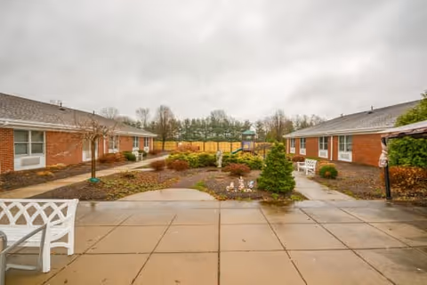 Outdoor courtyard area at St. Mary Villa for Independent & Retirement Living featuring paved walkways, garden beds with shrubs and small trees, white benches, and two single-story brick buildings on either side under a cloudy sky.