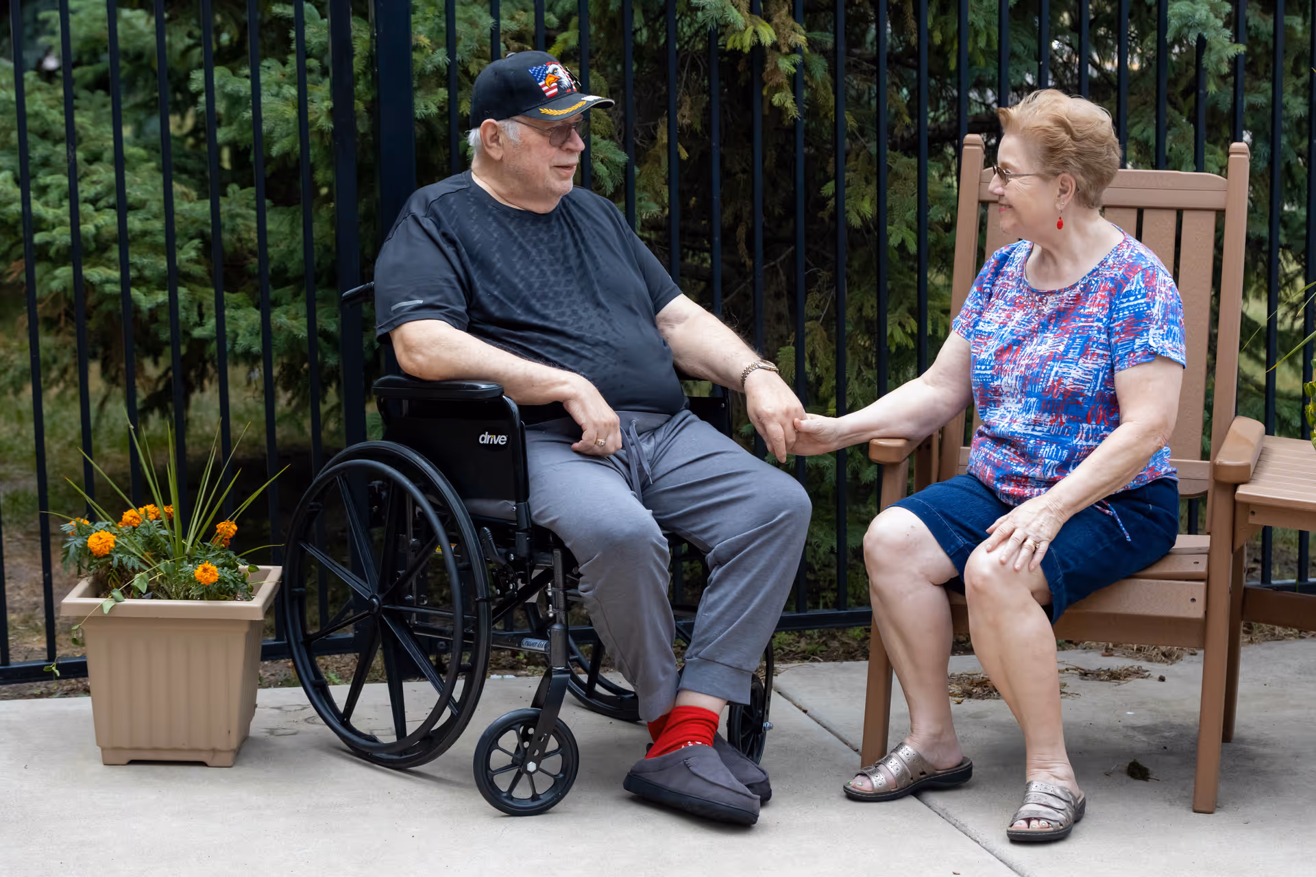 An elderly man in a wheelchair holding hands and smiling with an elderly woman sitting on a chair outdoors near a black metal fence and a planter with orange flowers.