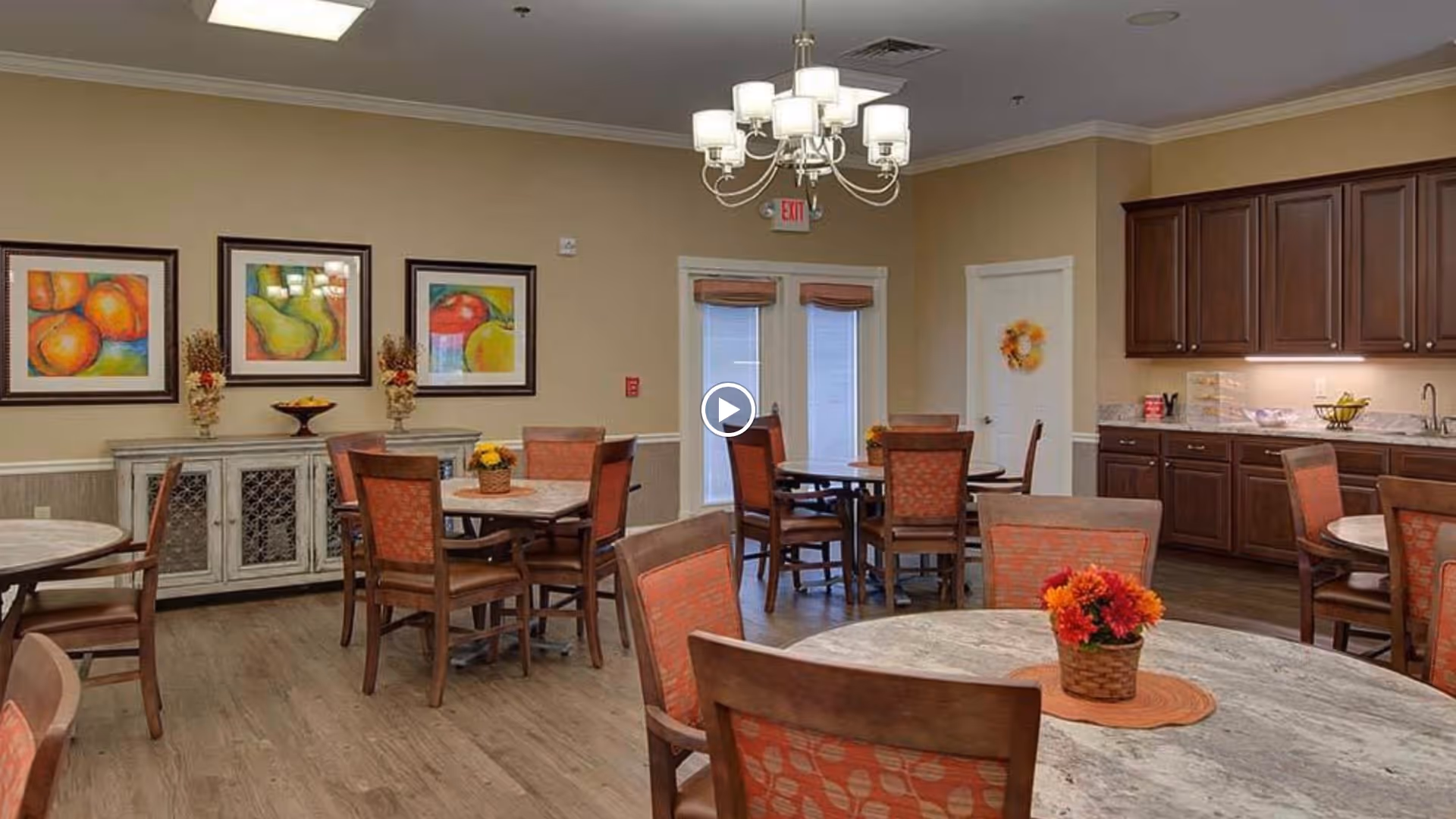 Communal dining room with round tables and chairs, fruit artwork on the walls, and a kitchenette along one side.